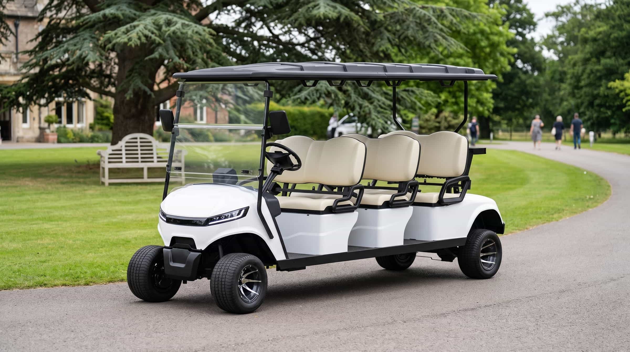 White six seater electric golf buggy with three forward facing benches on a drive beside the lawn of a country house hotel, a cedar and a garden bench behind