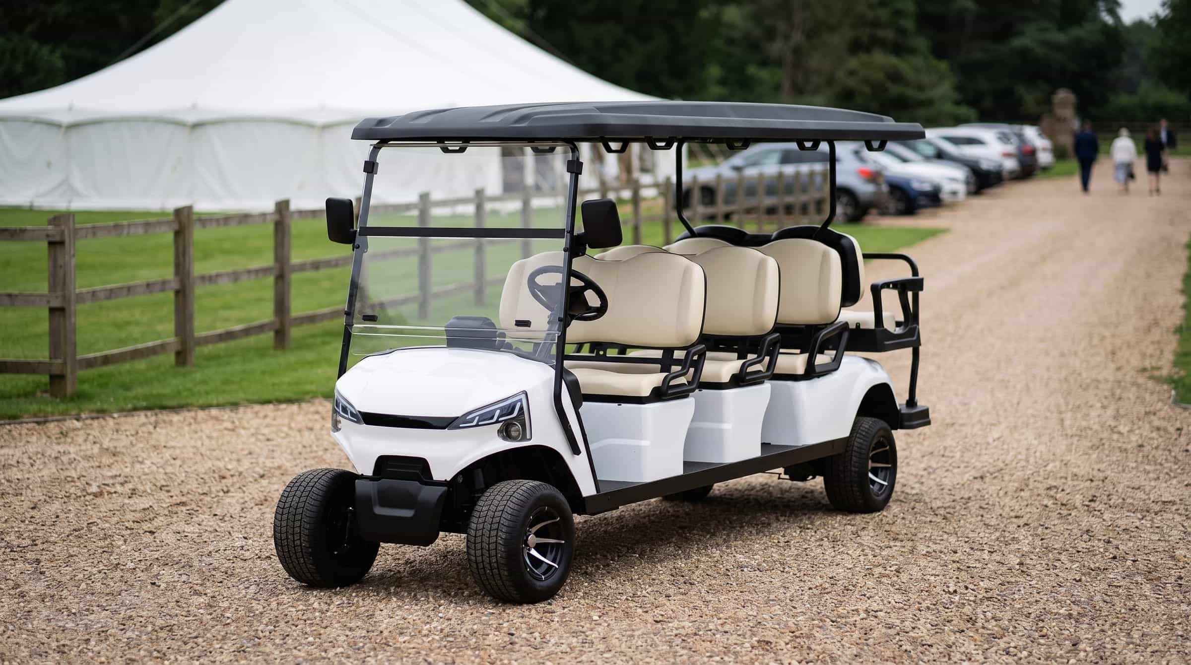 White eight seater electric golf buggy with three forward benches and a rear facing seat on the gravel drive of a country wedding venue, a marquee and parked cars behind