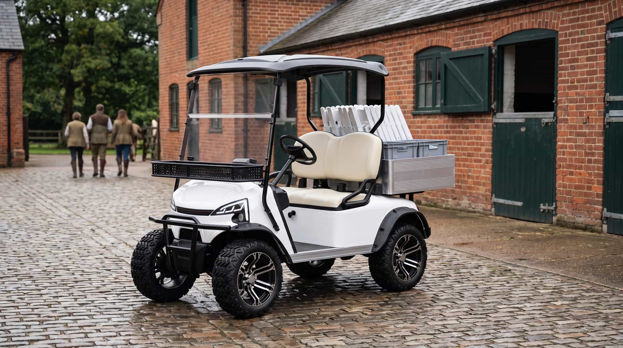 White two seat electric utility buggy on all terrain tyres, its aluminium load box stacked with folding chairs and crates, in the cobbled stable yard of a country estate