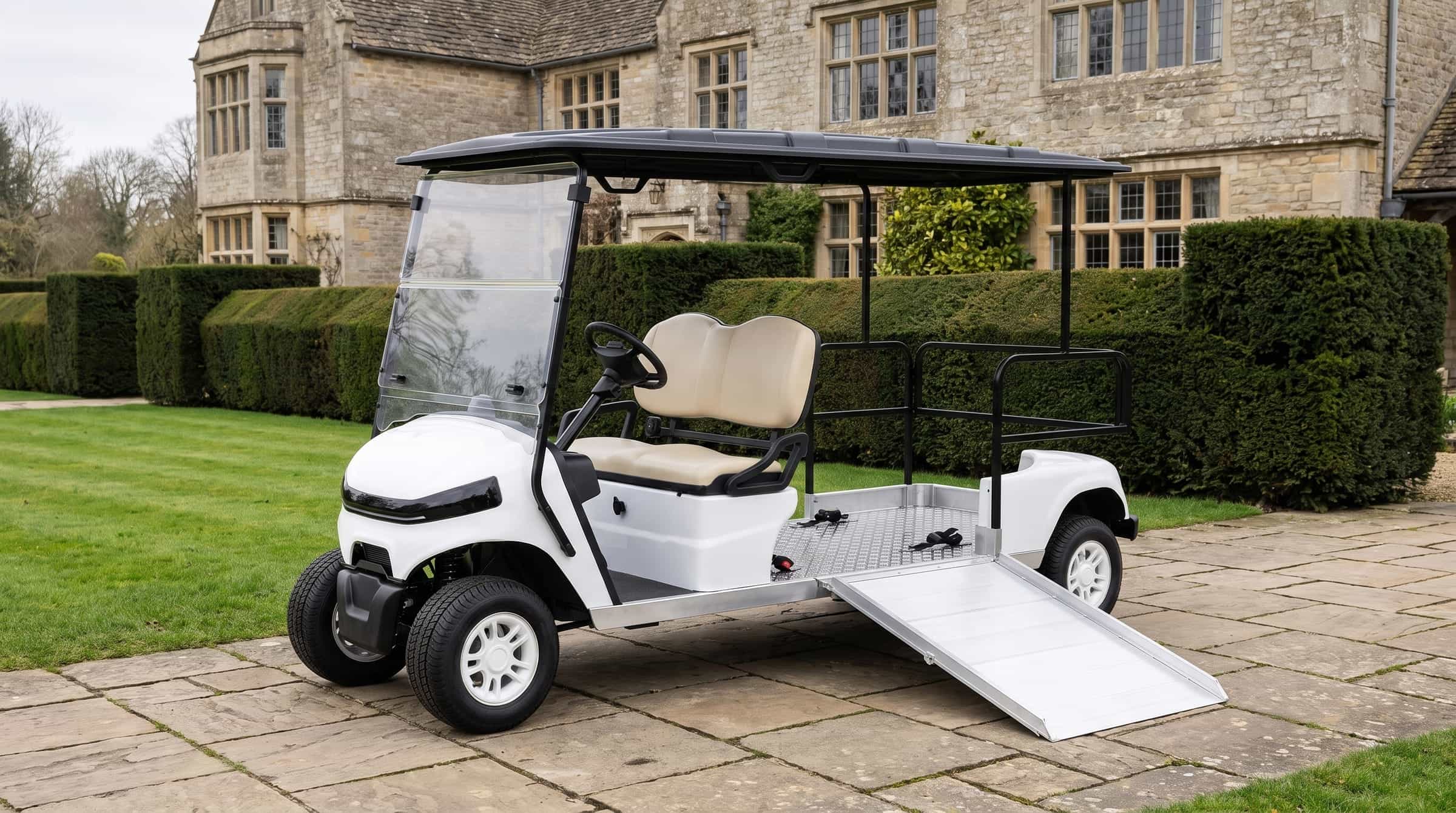 White electric buggy converted for wheelchair access, its aluminium side ramp lowered onto a stone terrace beside the flat wheelchair bay, a country house behind