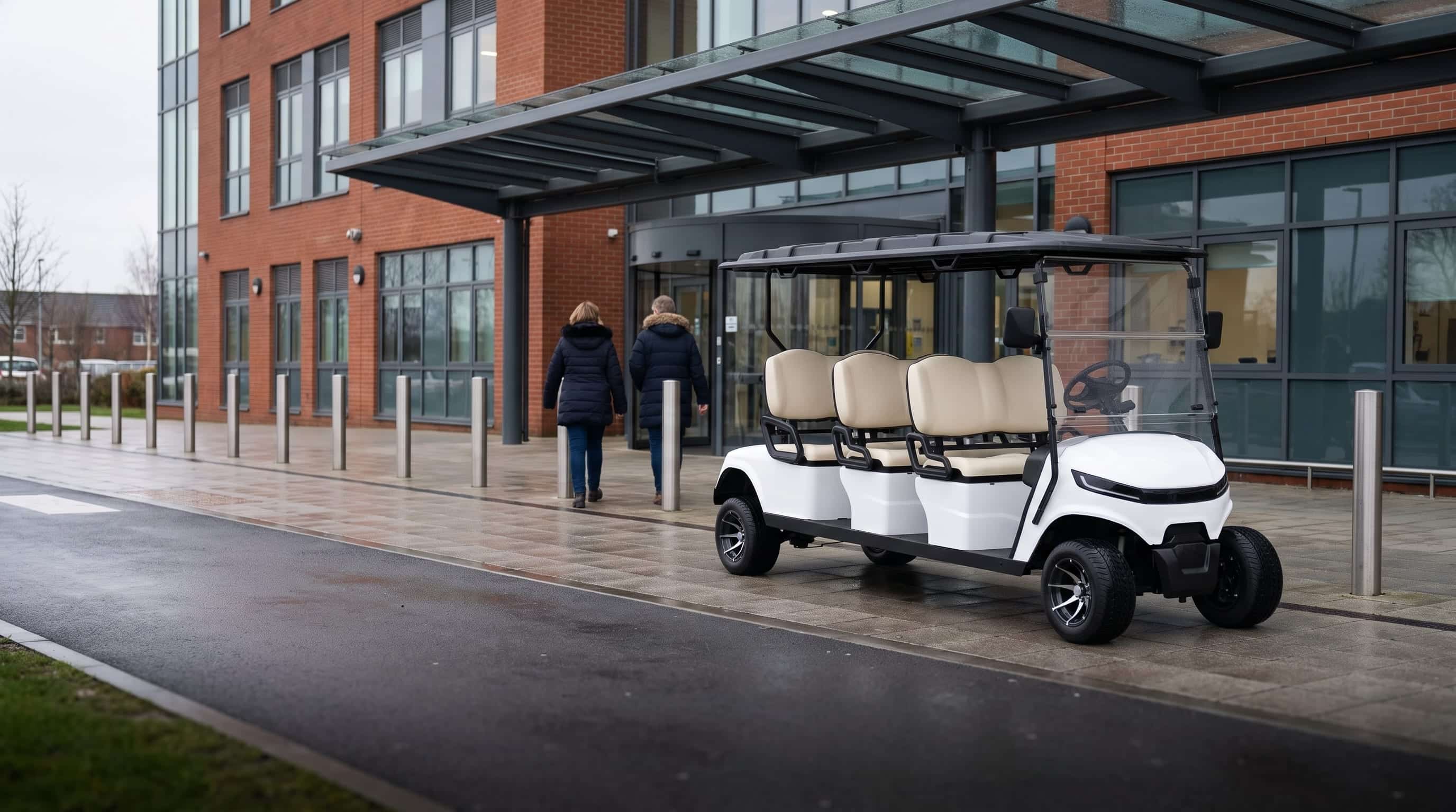 White six seater electric golf buggy waiting under the covered entrance of a modern hospital as two visitors walk in, on an overcast day