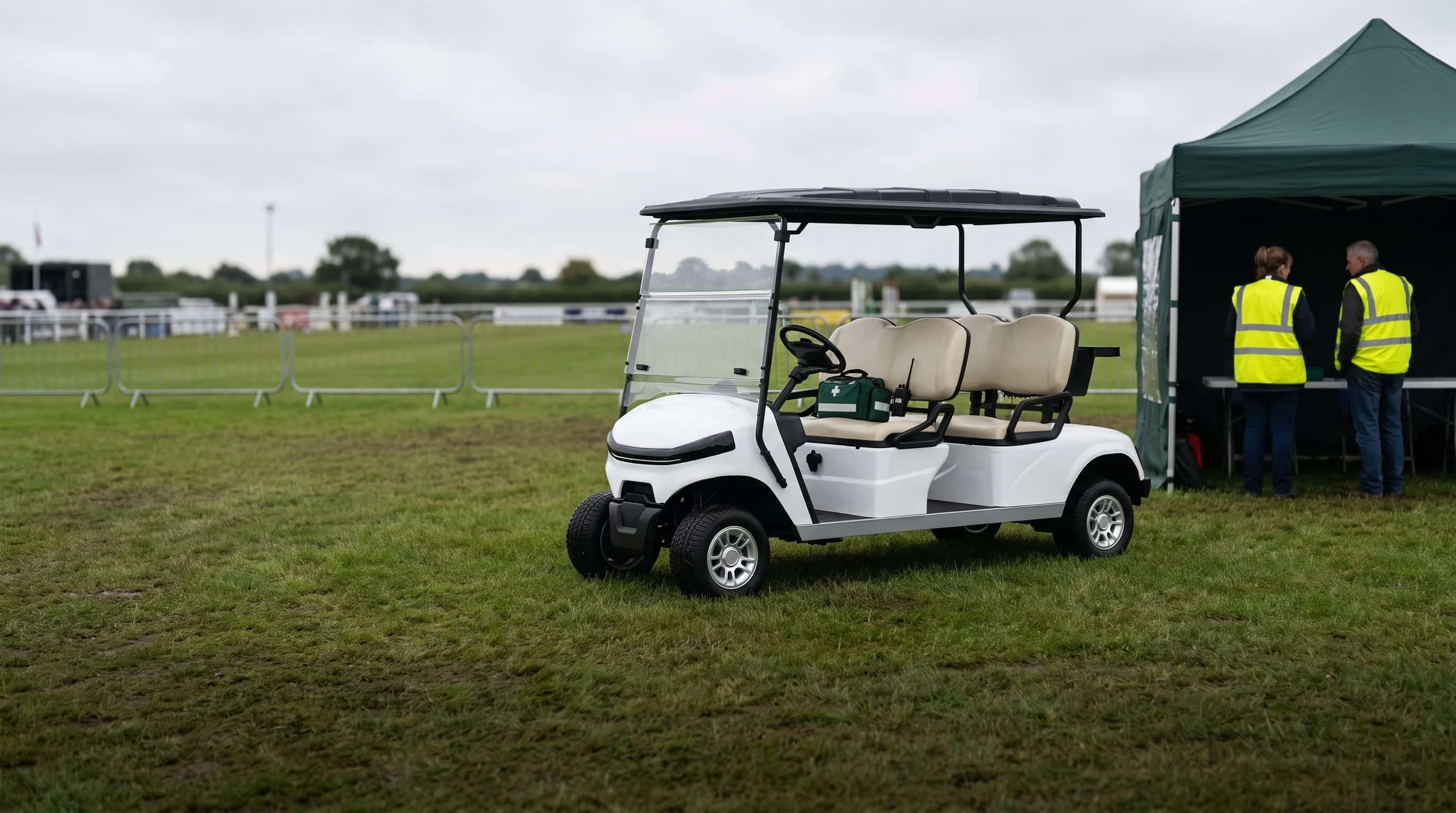 White four seater electric golf buggy with a first aid bag and a radio on the seat beside a stewards' gazebo at a showground arena, two stewards in high visibility vests